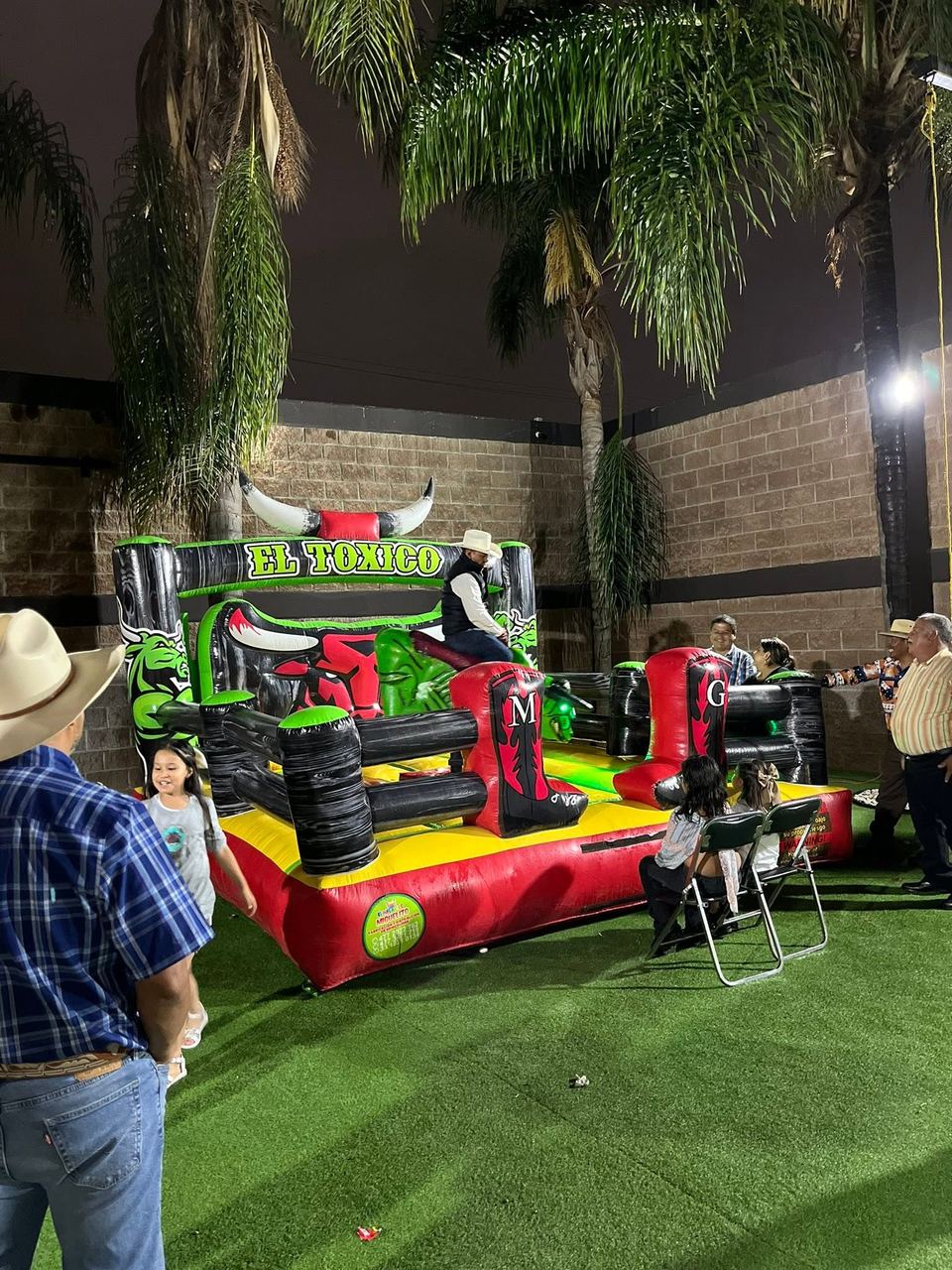 Toro mecánico verde El Tóxico montado en un jardín con palmeras durante un evento, con una persona de pie junto a la arena.